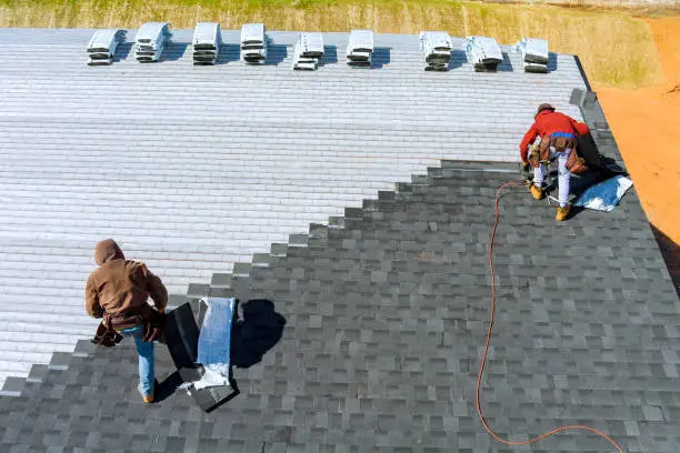 Two roofers installing dark gray architectural shingles over synthetic underlayment, with shingle bundles staged along the ridge, on a new roof in Washington, NC